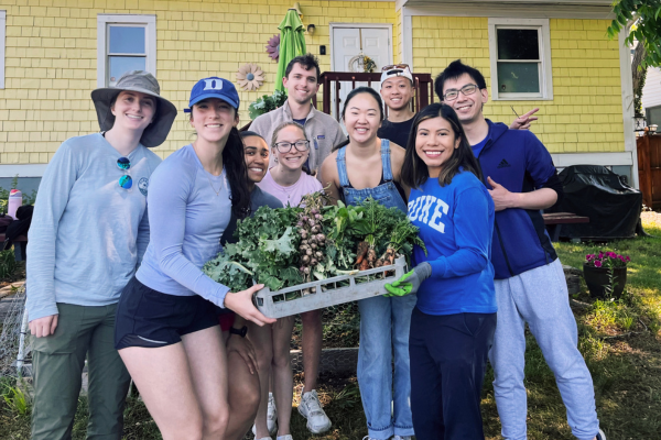 A group of eight people standing outdoors in front of a yellow house, smiling and holding a tray of freshly harvested vegetables.